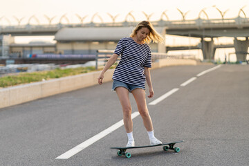 Active woman longboarder of middle age riding longboard on empty road or highway. Casual urban female relaxing on skateboard after work. Lady of 40s skateboarding at sunset. Modern lifestyle concept