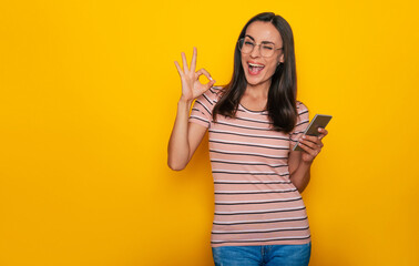 Modern beautiful young brunette woman is using smart phone for surfing on the internet and for work isolated on yellow background