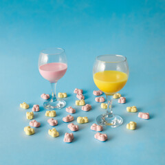 Two wine glasses with bright pink and yellow drink , surrounded with pink and yellow candy bears. Party arrangement. Pastel blue background. Minimal concept.
