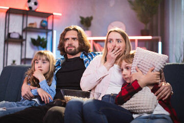 Caucasian family getting frightened and shocked while watching horror movie during evening time at home. Parents with two kids sitting on sofa with scary facial expressions.