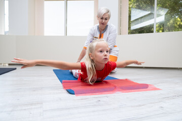 Little girl lying on fitness mat outstretching her arms to sides, doctor monitoring procedure execution, work with posture and ridge, rehabilitation centre