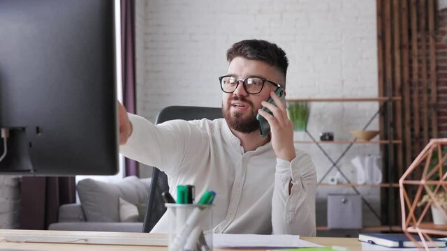 Portrait shot Handsome young man office worker in glasses speaking on the mobile phone and explaining something of working process while looking at the computer screen. Freelancer work in home office.