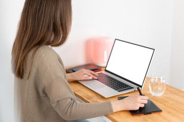 Unrecognizable brunette woman graphic designer in grey jacket working at the laptop and digital graphic tablet. Freelancer working at wooden desk from home office.
