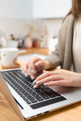 Close up photo of pretty woman hands typing on laptop keyboard. Female freelancer working from home. On the wooden desk notebook and cup of tea. Selective focus, blurred background. Side view.