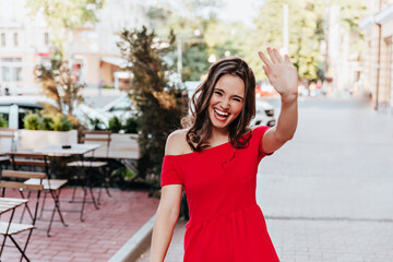 Happy woman in trendy red dress waving hand. Outdoor shot of positive beautiful girl walking by street cafe.
