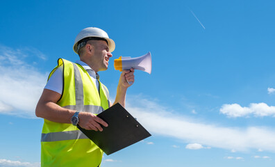 Builder, foreman or architect in a white helmet and a reflective vest shouts into a megaphone and holds a clipboard against a blue sky background, copy space, banner