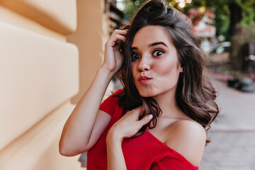 Cheerful dark-eyed lady making funny faces on the street. Elegant brunette girl in red dress joking during photoshoot.