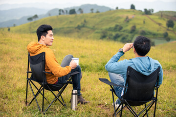 Asian man friends sitting on outdoor chair on the mountain drinking hot coffee together. Male...