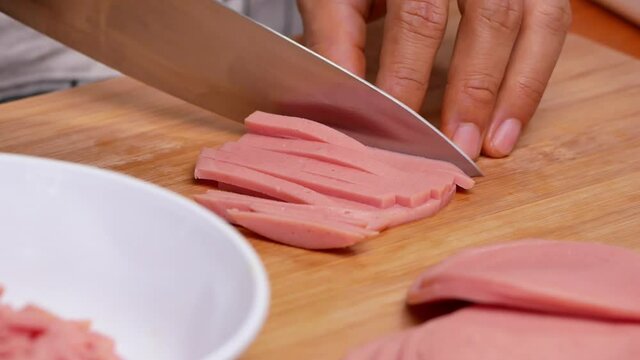Close-up of female hands cutting raw ham bologna into dice using a knife on the cutting board. Concept of preparing healthy meal.