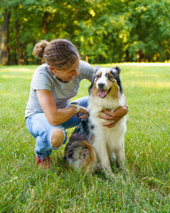 Naklejka na ściany i meble Happy 40s european woman sitting on green grass and hugging with young beautiful Australian Shepherd dog in meadow, positive female owner and cute purebred pet spending time together in nature