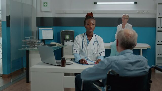 Patient with disability sitting at desk for checkup with medic at healthcare facility. Young doctor consulting elder man with handicap in wheelchair about treatment progress and healing