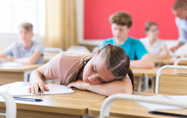 Tired teenage girl sleeping at desk in classroom during lesson with her head resting on her hands..