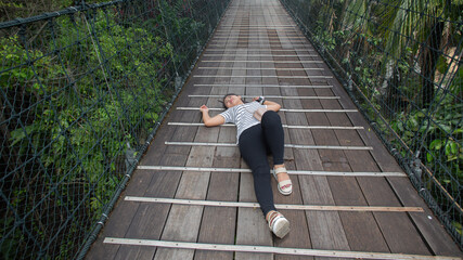 Young Malaysian woman laying on the wooden floor of a bridge surrounded by lush greenery in a park - Powered by Adobe