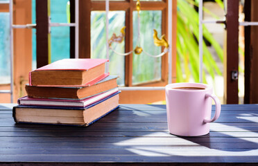 A cup of coffee is placed on the table by the window. with a pile of books and the morning sun shines in