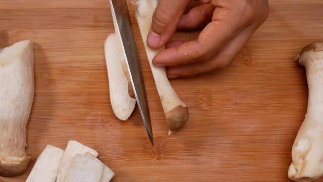 Close-up of female hands slicing oringi mushrooms or royal oyster mushroom using a knife on the cutting board. Concept of preparing healthy meal.