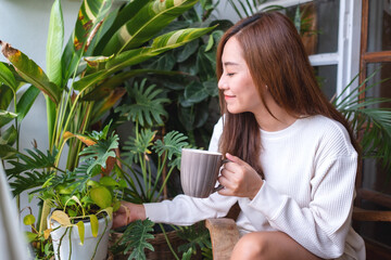 Portrait image of a beautiful asian woman drinking hot coffee and taking care of houseplants garden on balcony at home - Powered by Adobe