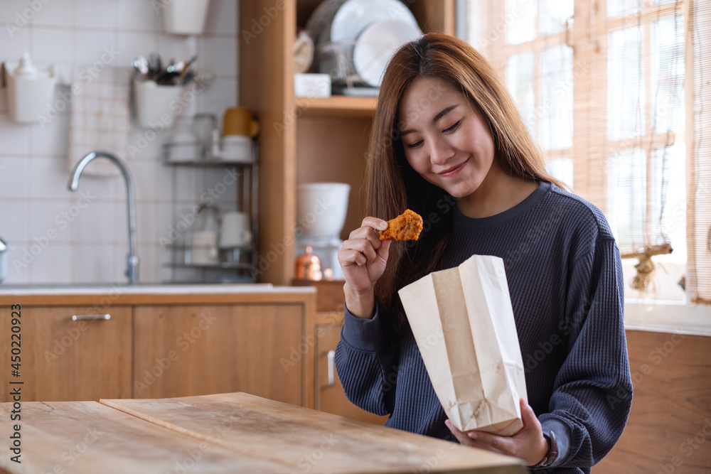 Wall mural portrait image of a beautiful young asian woman eating fried chicken from paper food bag in the kitc