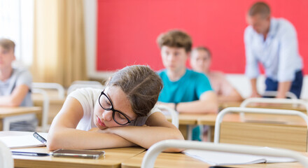 Fototapeta na wymiar Exhausted teen girl with glasses sleeping at desk in classroom during lesson on blurred background of classmates ..