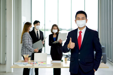 Asian businessman in a suit and face mask lifted a finger thumb up with a confident expression. The team is preparing for the meeting in the background. Everyone wears a mask. Concept of leadership.