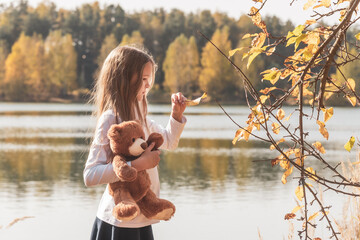 Pretty little girl in school uniform with cute teddy bear holds dry leaf on riverbank in sunny...