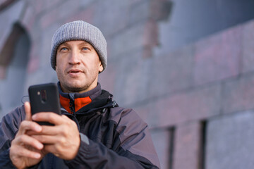 Portrait of happy smiling caucasian man with smartphone on city street against brick wall background. In an autumn jacket and hat. Using your phone in different ways