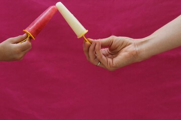 Child hand and a female hand holding white and pink ice cream bars on bright pink textured background with copy space