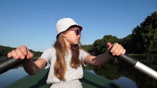 Little girl in sunglasses rowing along the river. Child controls the boat independently. Kid sails on a boat outdoors on summer vacation. Fishing with dad