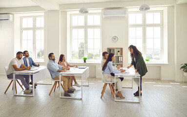Obraz premium Side view of successful people who are adult students and sitting at desks in a modern bright classroom. Female teacher who specializes in adult education teaches a variety of women and men.