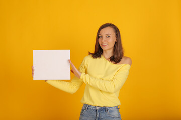 Naklejka na ściany i meble a young beautiful blonde girl in a yellow jacket and jeans holds a white sheet of paper in her hands
