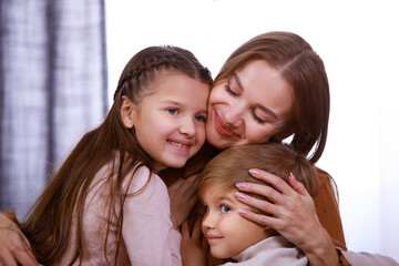 Portrait of a happy family in a room in daylight against the background of a window. Mom is enjoying the hugs of her adorable children, son and daughter. Happy family life. Close up.