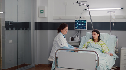 Obraz premium Specialist doctor putting oximeter checking pulse talking with sick woman explaining sickness symptom during recovery appointment. Patient resting in bed waiting for disease treatment in hospital ward
