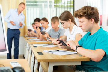 Focused teenager with group of fellow students sitting at lesson in classroom, using mobile phone..