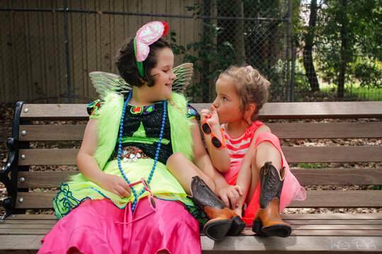 Two young girls wearing bright dress-up sitting on a park bench