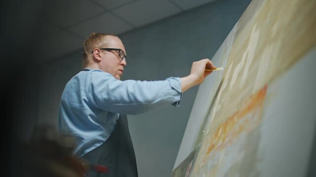 Artist's workshop, creative person at work, a talented adult man artist covers the canvas with beige oil paint, draws the modern abstract picture, 4k slow motion.