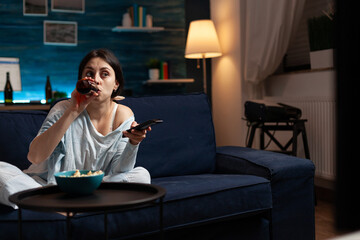 Confused young woman drinking beer while watching movie on tv changing channels using remote alone...