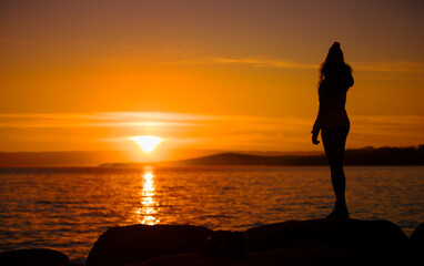 Silhouette of a young lady enjoying freedom by the sea at sunset. Slim relaxing woman in the evening looking away. Portrait Photography