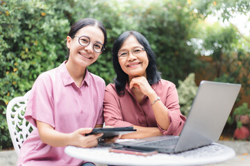 Young woman and senior mature wearing glasses using laptop and tablet at home. Smiling Asian family mother and daughter looking at camera spending time together with digital device.