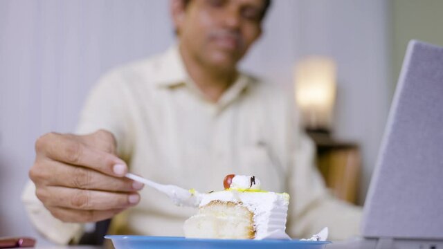 Rack focus from cake to person, Businessman eating cake while working on laptop at office - concept of unhealthy eating, sugar or diabetes and modern lifestyle workplaces