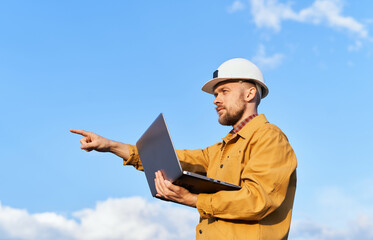 Cute bearded caucasian supervisor, architect, technician or building contractor in white hard hat and orange jacket using laptop and showing with hand. Blue sky with clouds background in sunny day