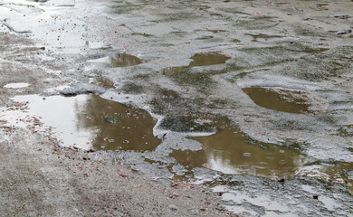 Fototapeta na wymiar A water-filled hole in an asphalt road. A rainy day in a big city, cars are driving along the old road. Bad road with asphalt in pits and potholes that are dangerous for motorists and pedestrians.