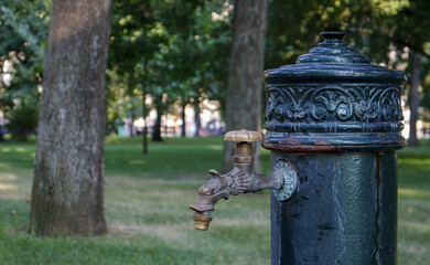 Fototapeta na wymiar Water well in the park in summer, pumping system, close-up and side view of a beautiful metal bronze faucet. Part of an old iron outdoor tap. Click on the mineral water pump room.