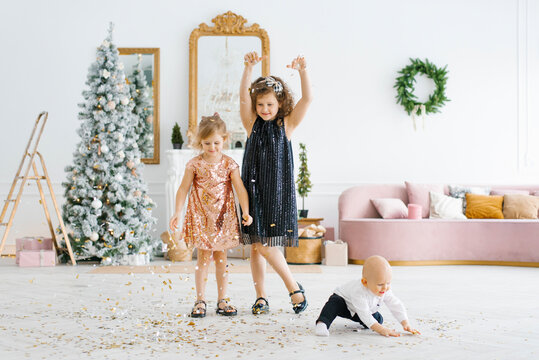 Two girls and a little boy play with Christmas confetti in the living room near the Christmas tree