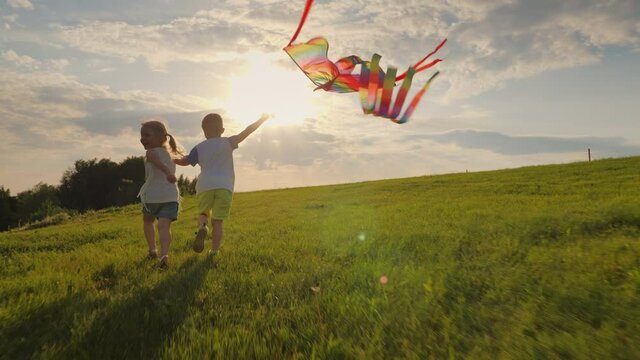 Happy smiling little children running holding hands and playing with a kite on the meadow in the rays of the sun at sunset