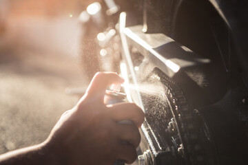 Outdoor sun reflect photograph of hand applying spray oil on motorbike chain for maintenance.