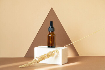 Mock-up of glass dropper bottle with dry sprig of grass, standing on wooden geometric stand in rays...