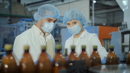 Two specialists, a young man and a middle-aged woman, in white coats, disposable hats and medical masks, are discussing production issues against the backdrop of plastic bottles.