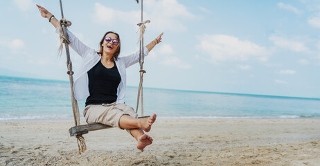 Happy adult young woman in sunglasses and white blouse swinging on a swing on a tropical beach - obrazy, fototapety, plakaty