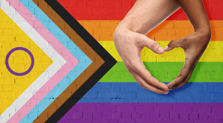 lgbtq, trans and intersex rights concept - close up of couple wearing rainbow ribbon wristbands and showing hand heart over progress pride flag on background - obrazy, fototapety, plakaty