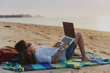 Young woman freelance digital nomad lying on the beach with laptop at sunset