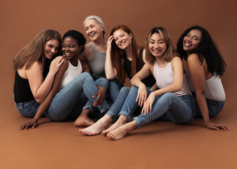 Six women of different ages sitting together in studio on brown background. Multi-ethnic group of diverse females having good times. - Powered by Adobe